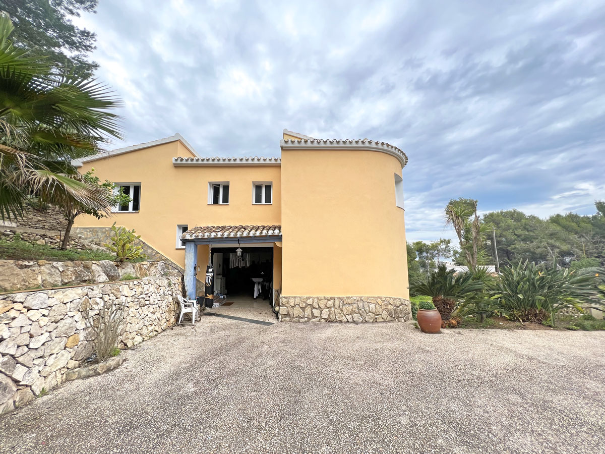 Fachada exterior de estilo mediterráneo con muros de piedra natural, zona de aparcamiento en grava y vegetación autóctona rodeando la entrada.