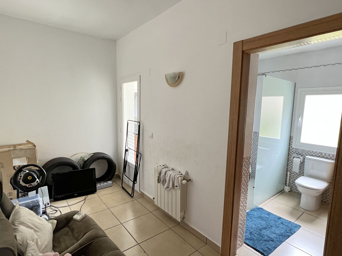 Habitación funcional con suelos de gres y radiador, conectada a un baño privado con ducha de obra, mampara de cristal y ventilación natural.
