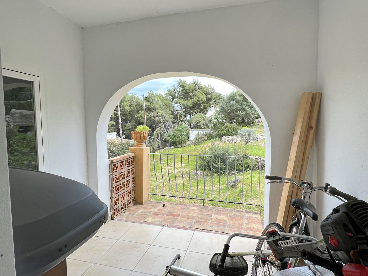 Porche cubierto con arco mediterráneo y vistas a la zona verde. Espacio funcional con suelos cerámicos, barandilla metálica y detalles en terracota.