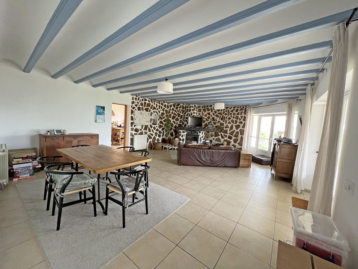 Salón-comedor diáfano con vigas de madera a la vista en azul claro, pared frontal revestida de piedra natural y suelos de gres cerámico en tonos neutros.
