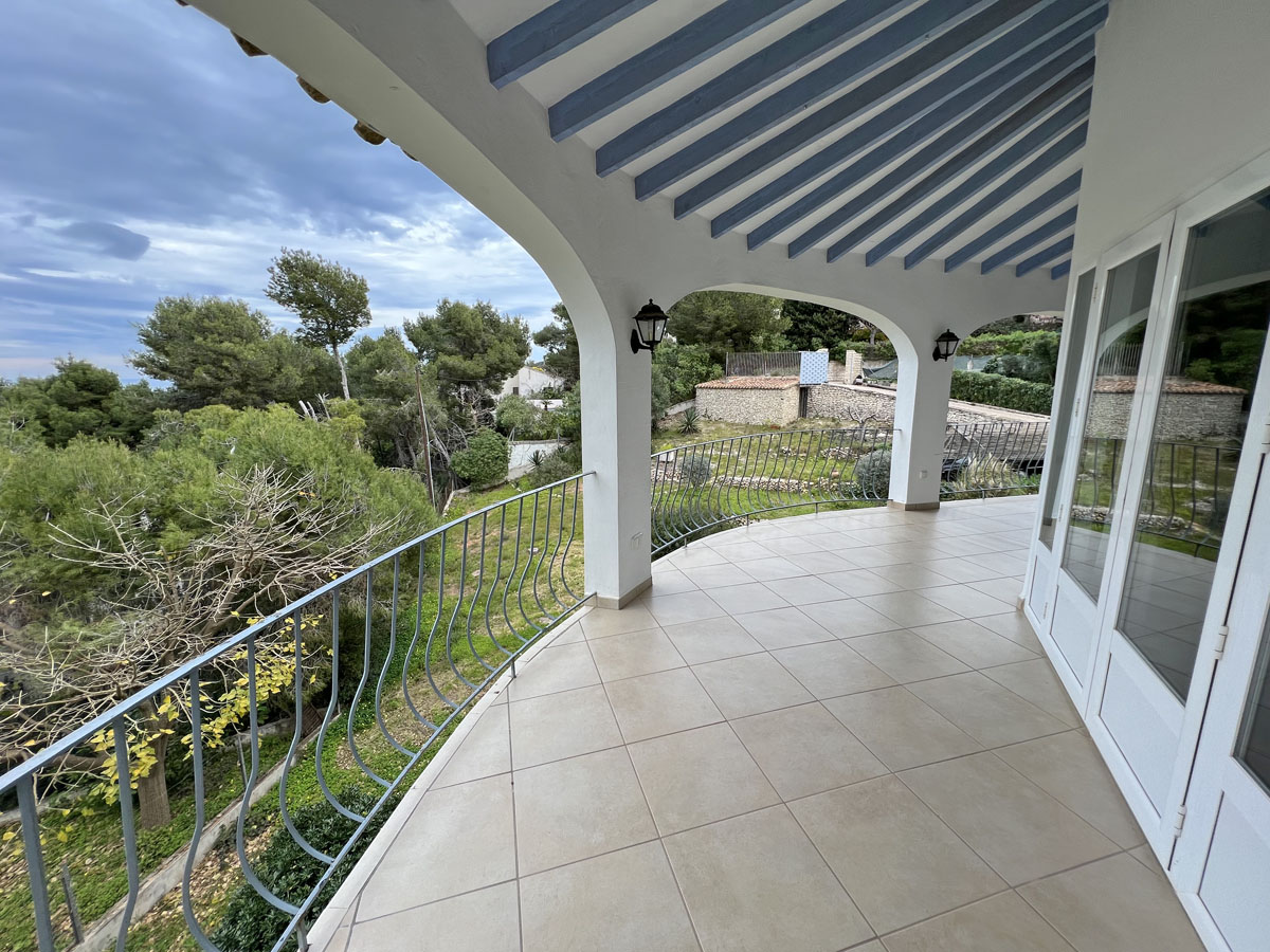 Terraza cubierta con arcos blancos, vigas de madera azules y suelo cerámico, con vistas al jardín mediterráneo y al horizonte marino.