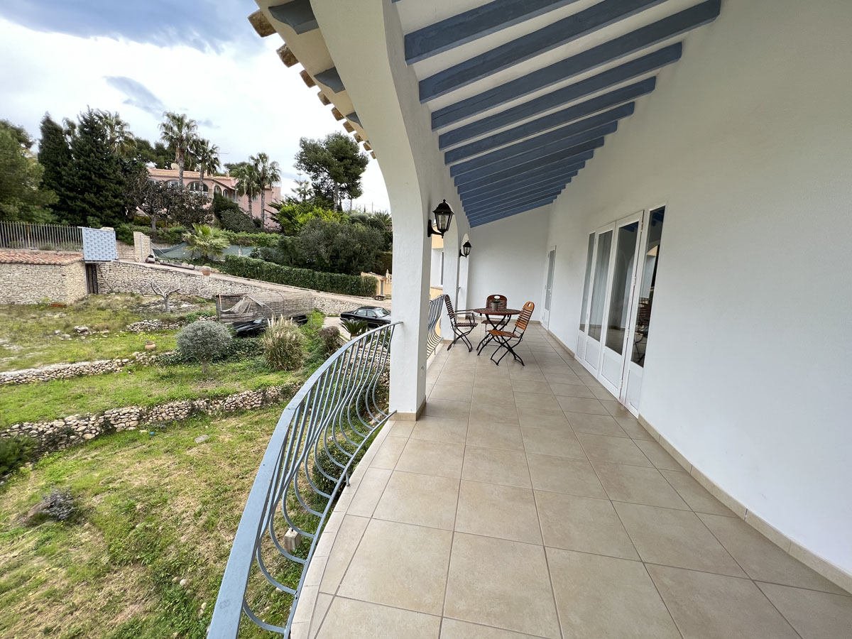 Terraza cubierta con vigas de madera en azul, suelos de gres y barandilla curva metálica con vistas al jardín abancalado y muros de piedra natural.