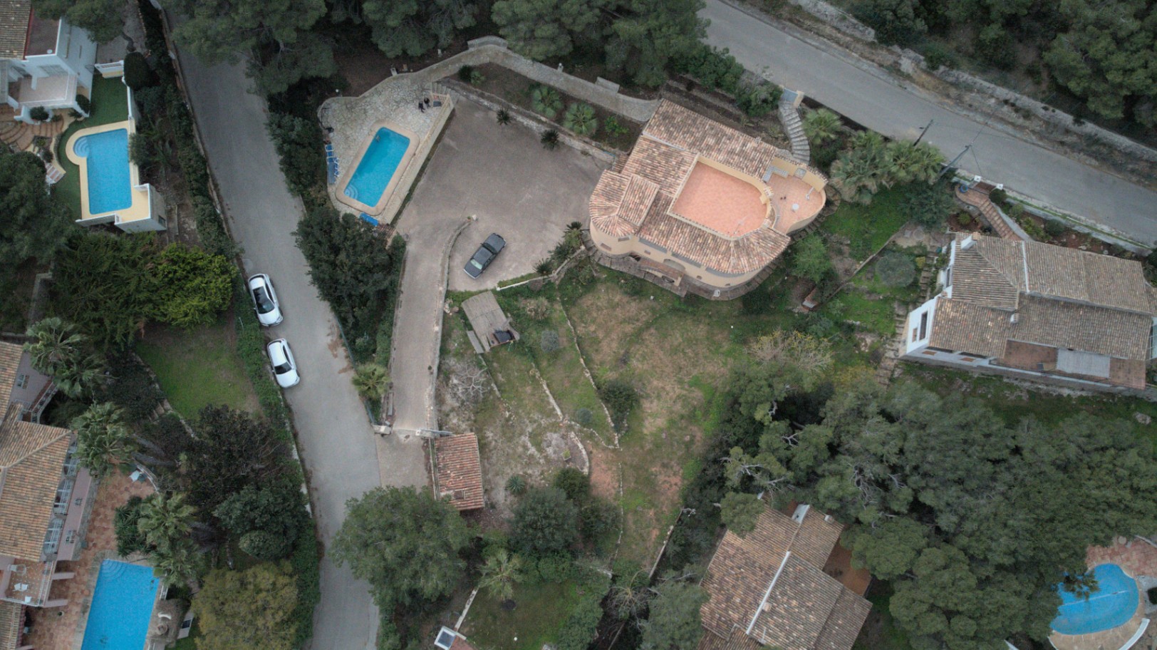 Vista aérea de la propiedad que muestra la distribución de la villa, la amplia zona de aparcamiento pavimentada, la piscina privada y el terreno aterrazado rodeado de pinos.