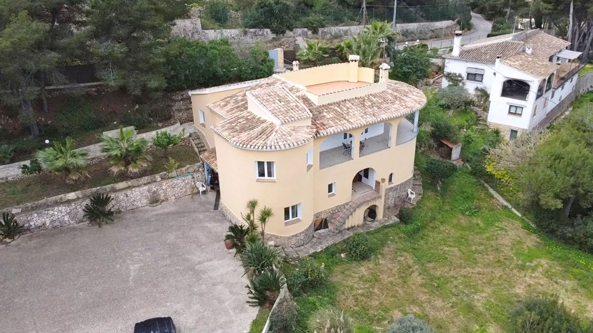 Vista aérea de la villa con fachada curva, base de piedra natural y cubiertas de teja cerámica, rodeada de jardines mediterráneos y amplia zona de aparcamiento.