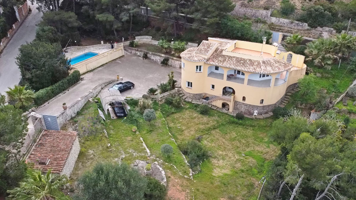 Vista aérea de la villa con fachada curva y base de piedra, destacando la amplia zona de aparcamiento, la piscina privada y el jardín mediterráneo en una parcela de 1.377 m².