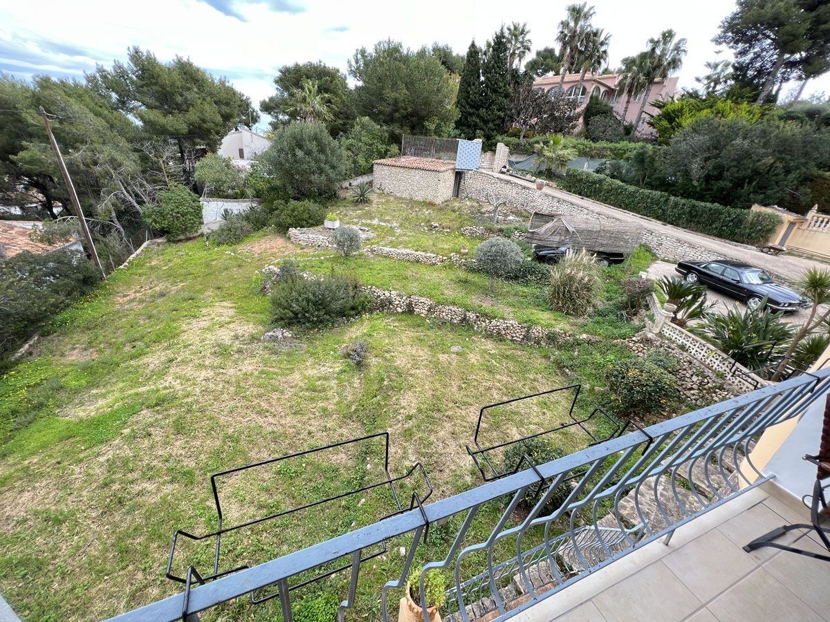 Vista desde el balcón hacia la amplia parcela aterrazada con muros de piedra natural, vegetación mediterránea y zona de aparcamiento lateral.