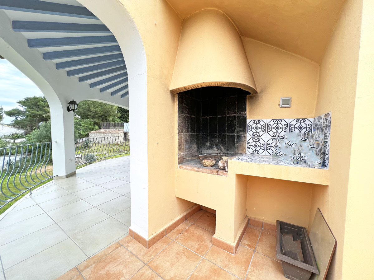 Zona de barbacoa de obra en la terraza, con encimera decorada con azulejos tradicionales, campana integrada y vistas al jardín desde el porche arqueado.