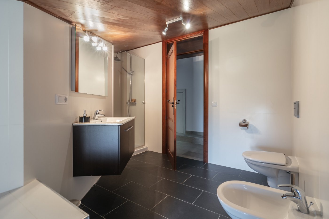 Baño moderno con techo de paneles de madera, suelo de baldosas oscuras, lavabo suspendido y ducha con mampara de cristal y grifería cromada.