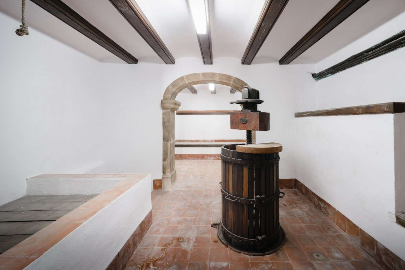 Bodega tradicional con prensa de madera antigua, arco de piedra tosca, vigas de madera a la vista y suelos de terracota.