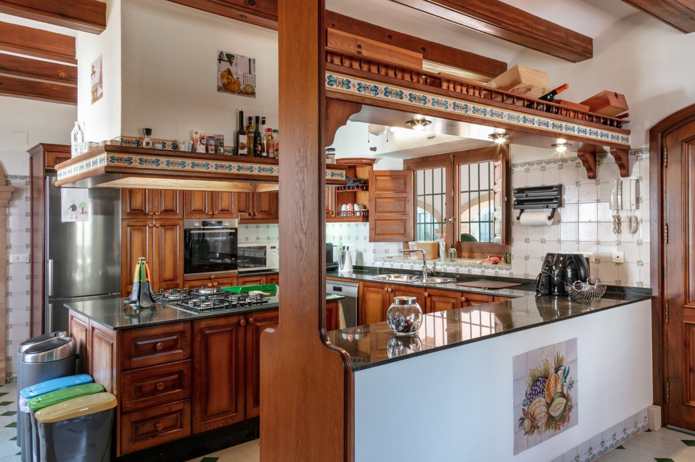 Cocina de estilo rústico con vigas de madera, encimeras de granito oscuro y azulejos tradicionales. Dispone de isla con fogones y electrodomésticos integrados.