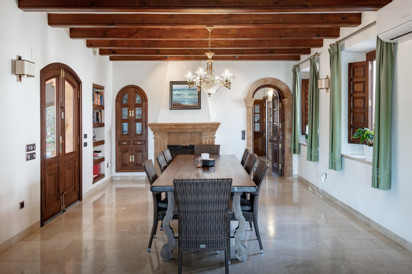 Comedor con vigas de madera, chimenea de piedra tosca y suelos de mármol. La estancia incluye arcos de piedra natural y una amplia mesa con sillas de mimbre.
