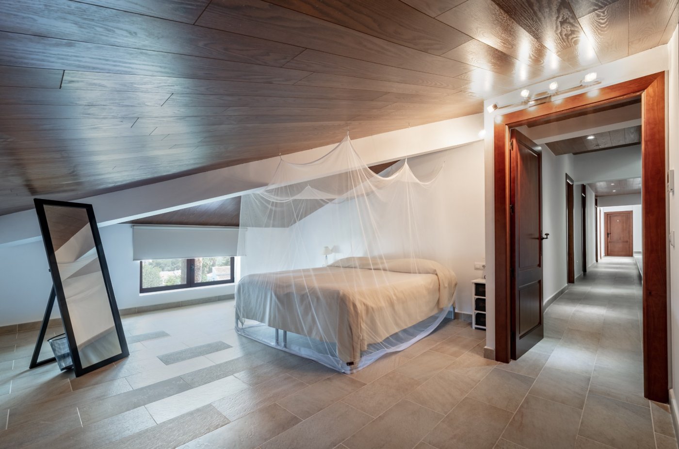 Dormitorio abuhardillado con techos de madera oscura, suelos de gres y cama con mosquitera. Un pasillo con marcos de madera maciza conecta con el resto de la planta superior.