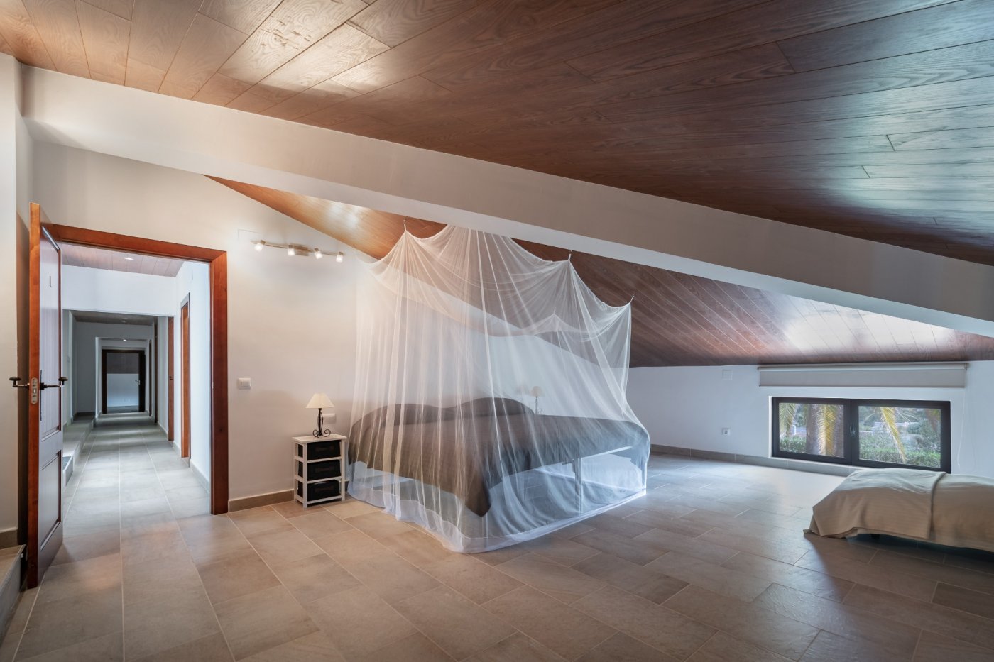 Dormitorio abuhardillado con techos de madera, suelos cerámicos y cama con mosquitera. Incluye ventana baja con vistas al jardín y acceso directo al pasillo.