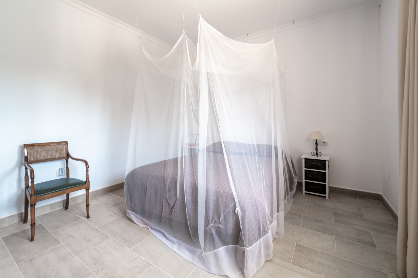 Dormitorio con cama doble protegida por una amplia mosquitera, suelos de gres cerámico y mobiliario auxiliar de estilo clásico.