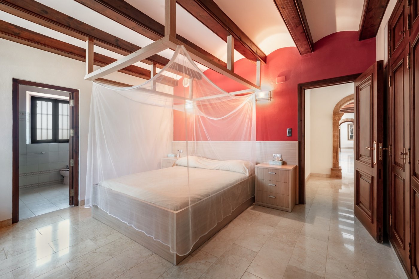 Dormitorio con vigas de madera, cama con dosel y mosquitera, pared roja y suelos de mármol, con acceso a baño privado y pasillo con arcos de piedra tosca.