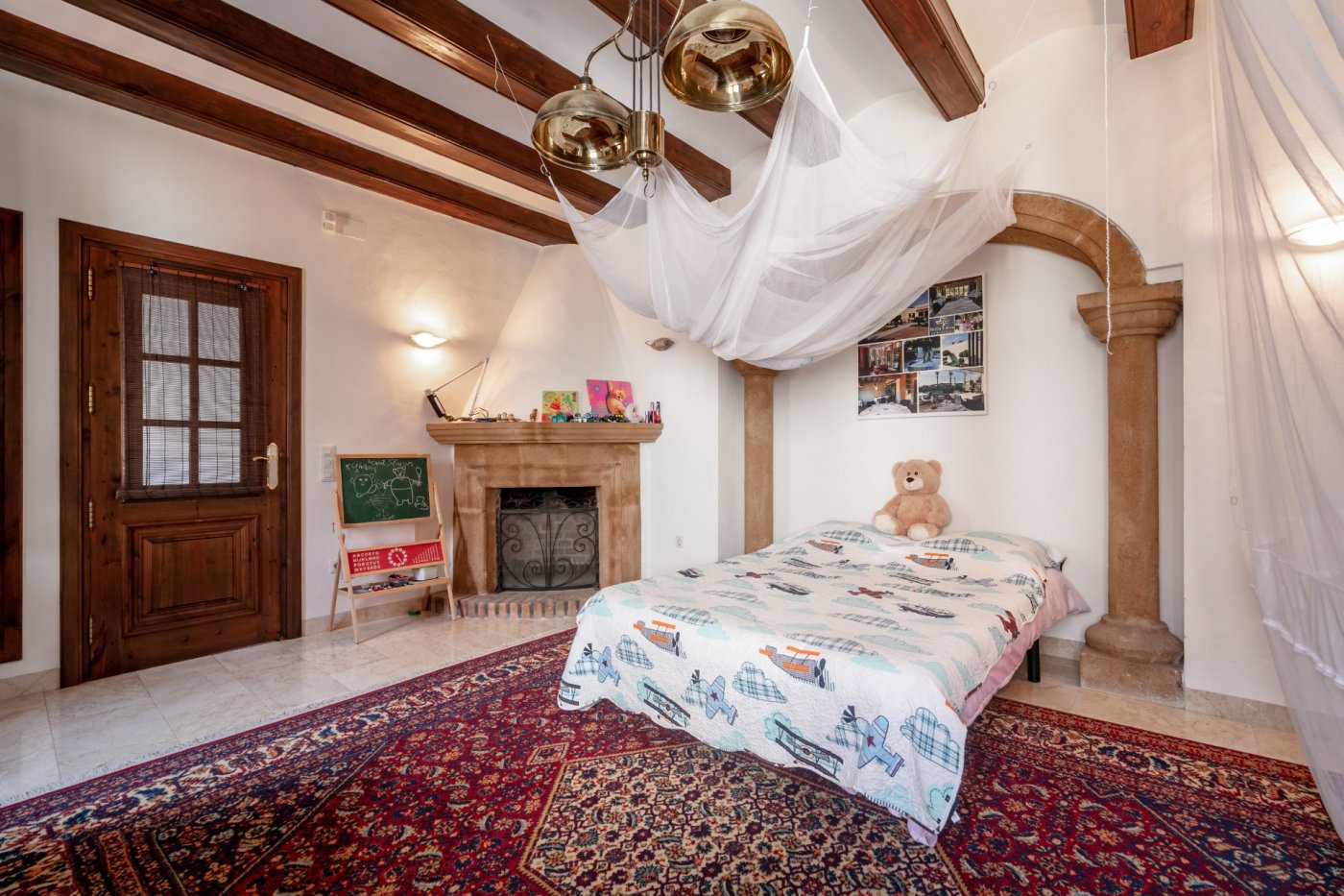 Dormitorio con vigas de madera, chimenea de piedra tosca y un arco tradicional sobre columnas clásicas que enmarca la zona de descanso.