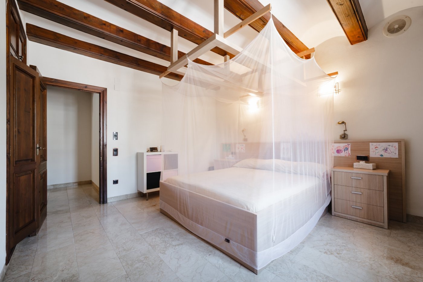 Dormitorio con vigas de madera vista, cama con dosel de mosquitera y suelos de mármol. Incluye puertas de madera maciza y una iluminación cálida integrada.