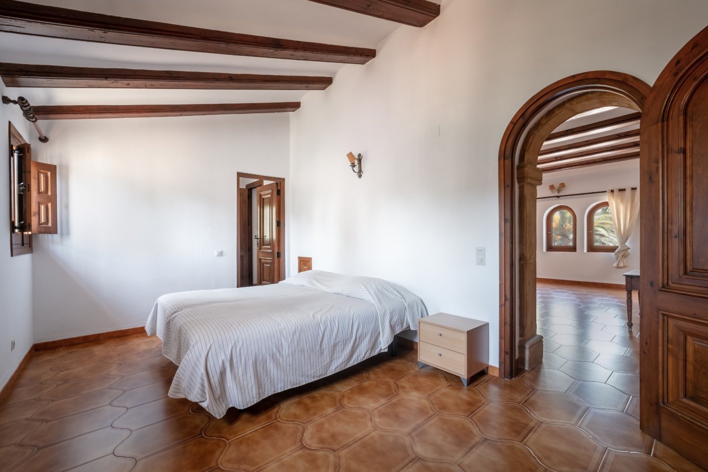 Dormitorio con vigas de madera vista, suelos cerámicos tradicionales y un gran arco de piedra tosca que conecta con una estancia contigua luminosa.