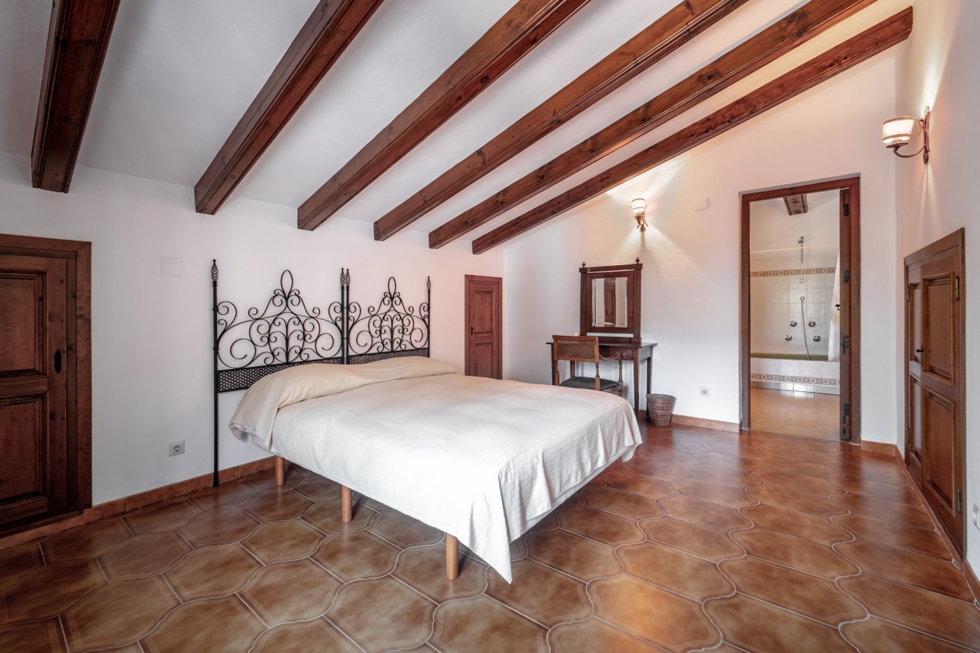 Dormitorio de estilo mediterráneo con techos abuhardillados de vigas de madera, suelos de gres rústico, cabecero de forja y acceso directo a baño en suite.
