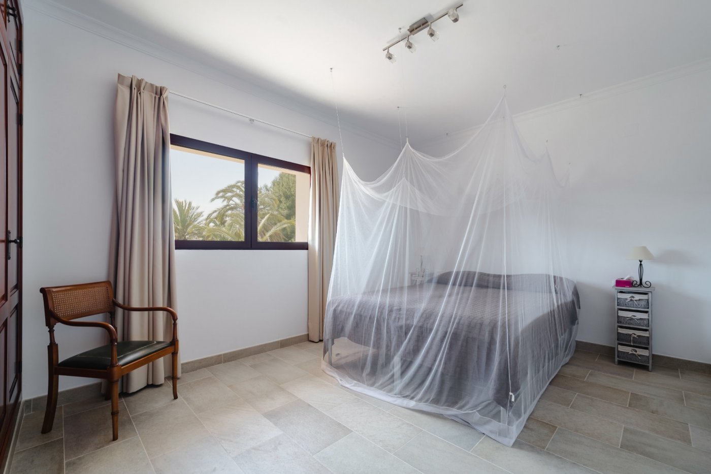 Dormitorio luminoso con cama de matrimonio y mosquitera suspendida, suelos de gres cerámico y ventana con vistas a las palmeras del jardín.