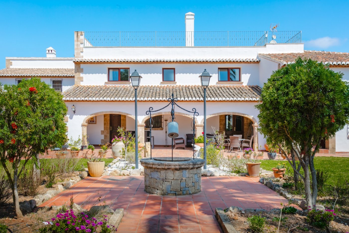 Patio exterior con pozo de piedra tradicional, rodeado de suelos de barro y una fachada blanca con arcos de piedra tosca y techos de teja mediterránea.
