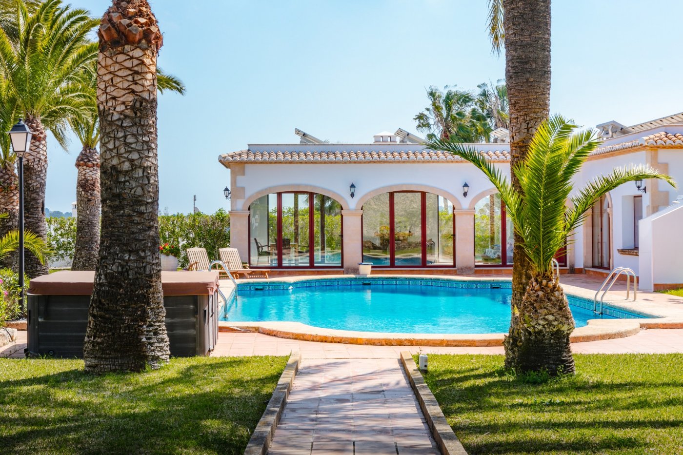 Piscina exterior rodeada de palmeras y césped, junto a una fachada con arcos de piedra tosca, ventanales de madera y placas solares en el tejado.