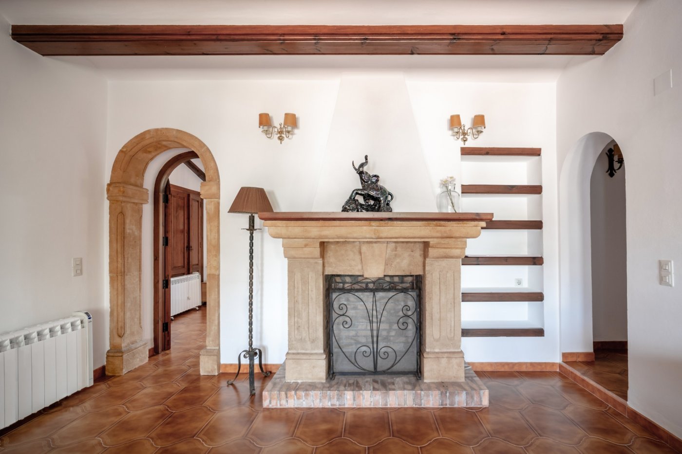 Salón con chimenea de piedra tosca, suelos de terracota y vigas de madera. Destaca el arco de piedra natural y las estanterías de obra integradas en la pared blanca.