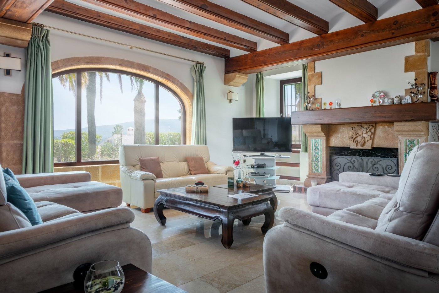 Salón de estilo mediterráneo con vigas de madera, gran ventanal en arco de piedra tosca y chimenea rústica con vistas al jardín y al valle.