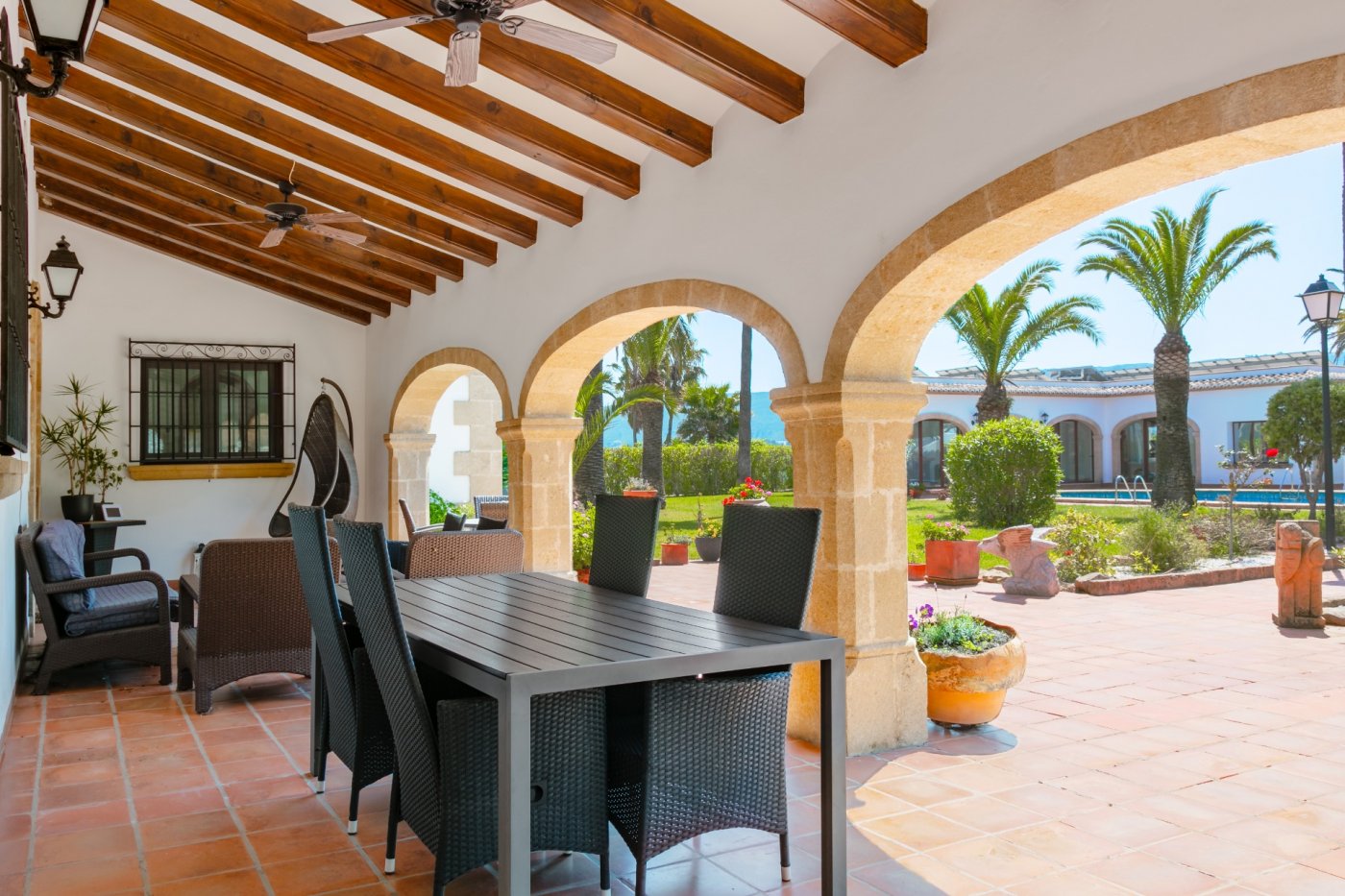 Terraza cubierta con arcos de piedra Tosca y vigas de madera, equipada con zona de comedor y estar frente al jardín con palmeras y piscina.