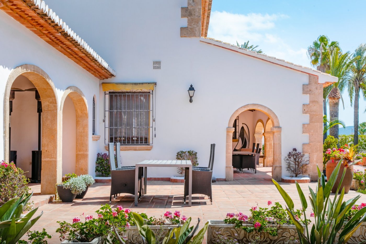 Terraza exterior con suelo de barro cocido, arcos de piedra Tosca y zona de comedor. La fachada blanca resalta entre la vegetación mediterránea y las palmeras.
