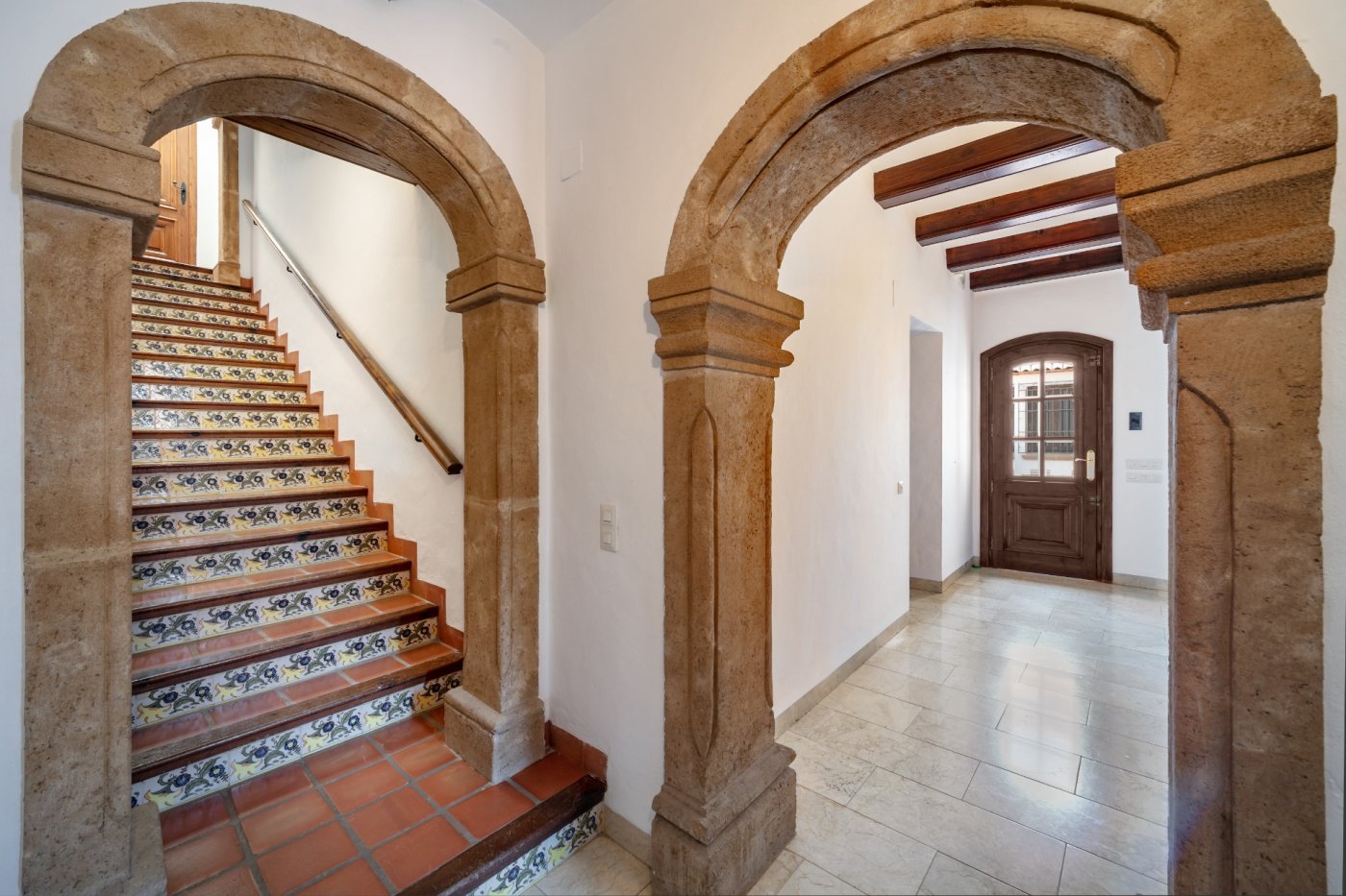 Vestíbulo con arcos de piedra tosca que dan paso a una escalera con contrahuellas de azulejo decorativo y un pasillo con vigas de madera vistas.