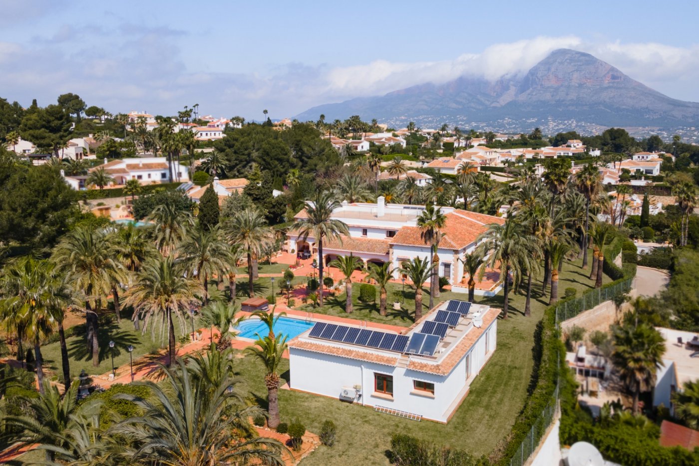 Vista aérea de la finca con villa principal, casa de invitados equipada con placas solares, piscina y extenso jardín de palmeras con el Montgó al fondo.