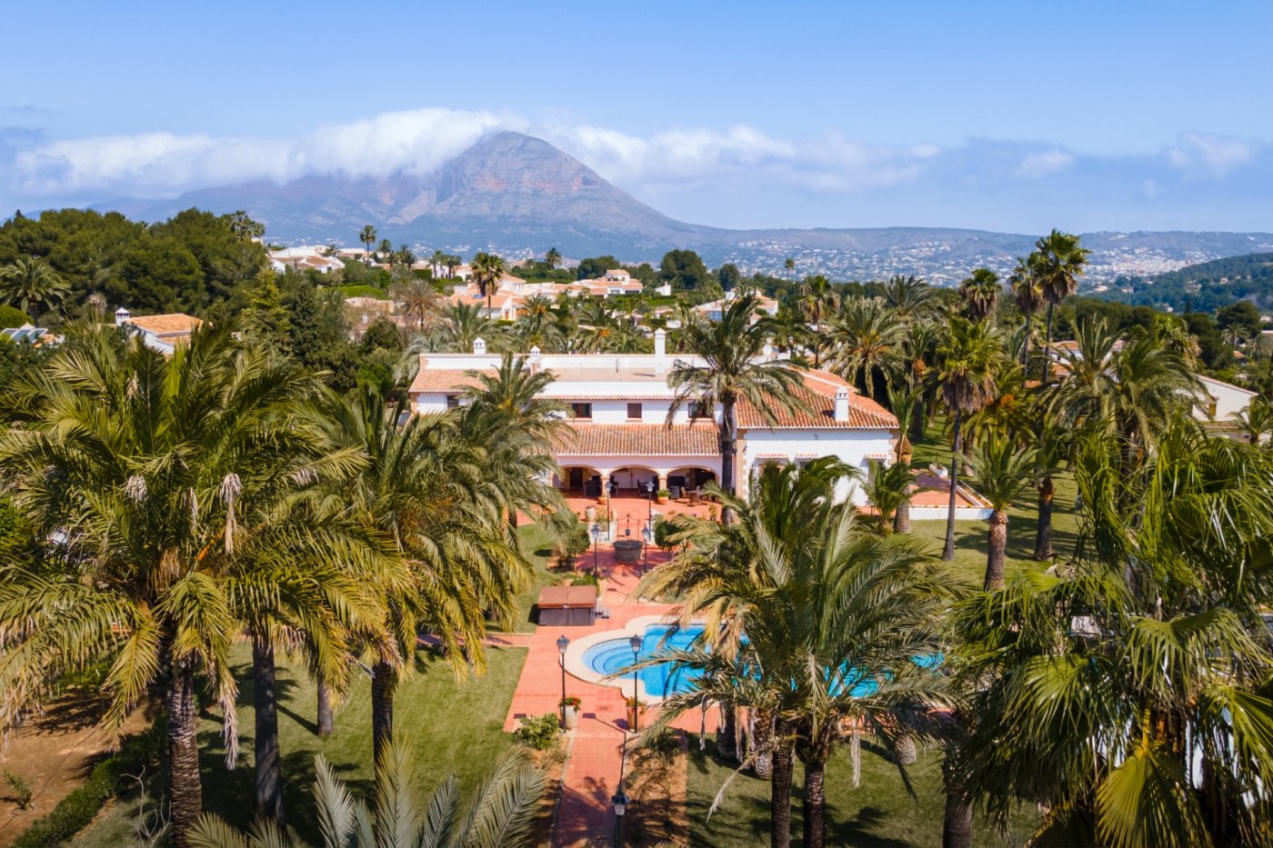 Vista aérea de la villa mediterránea con su extensa parcela de palmeras, piscina privada y el emblemático monte Montgó al fondo.