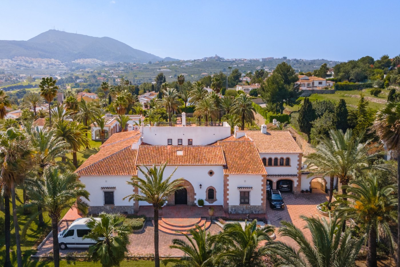 Vista aérea de la villa mediterránea con tejados de teja, arcos de piedra tosca y una amplia parcela rodeada de palmeras con vistas panorámicas al valle.