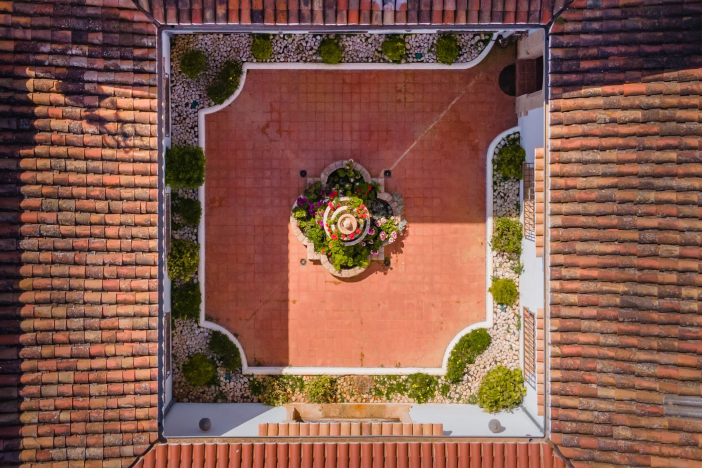 Vista cenital del patio interior central con suelo de terracota, una fuente ornamental con flores y jardineras perimetrales rodeadas por tejados tradicionales.