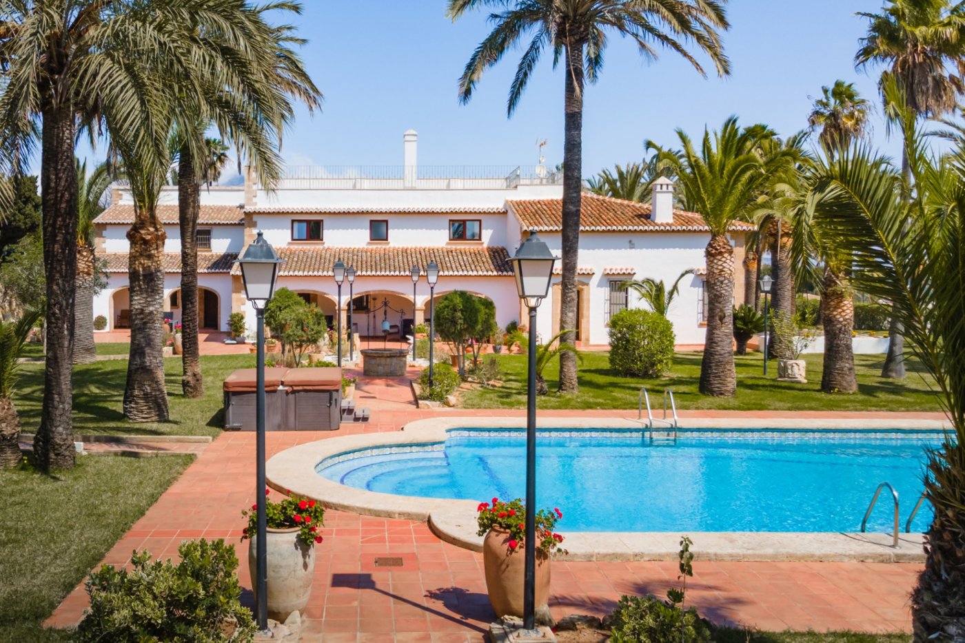 Vista exterior de la villa mediterránea con piscina, rodeada de un jardín con palmeras, césped y una amplia terraza de terracota con farolas decorativas.