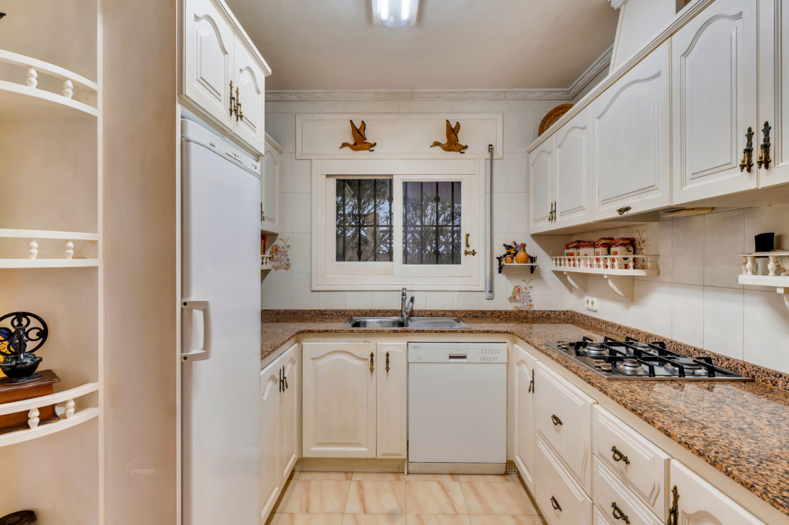 Cocina equipada con armarios blancos de estilo clásico, encimera de granito, placa de gas y lavavajillas integrado bajo una ventana con luz natural.