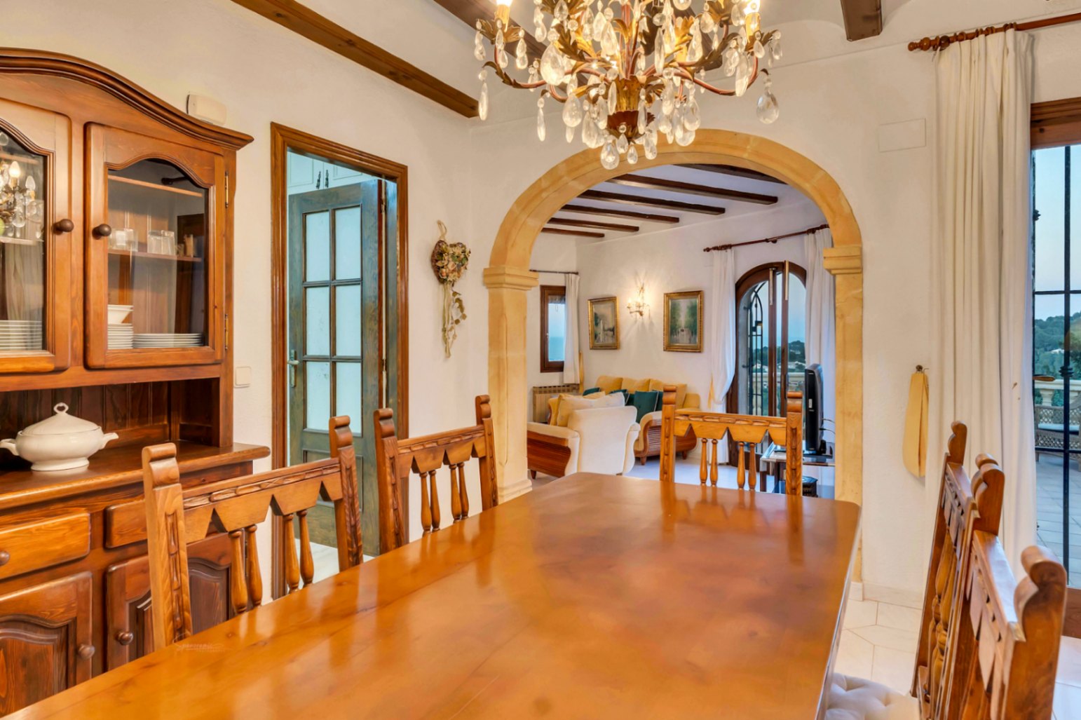 Comedor tradicional con mesa de madera maciza, alacena rústica y arco de piedra tosca. Destacan las vigas expuestas y la lámpara de cristal que conecta con el salón.