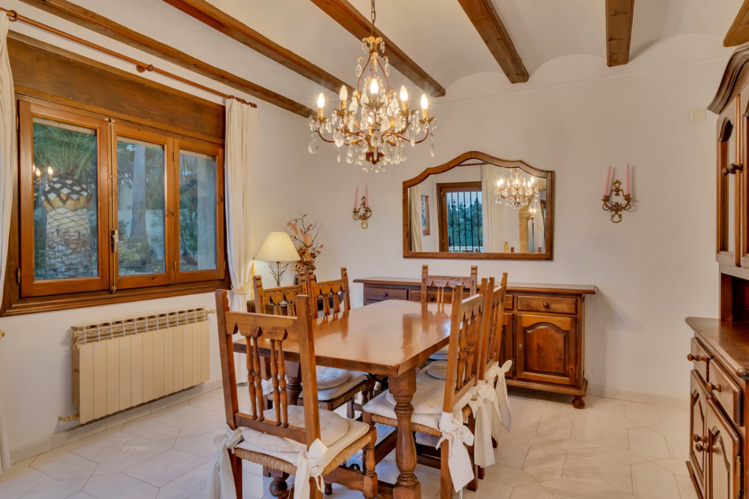 Comedor tradicional con vigas de madera, mesa de madera maciza, lámpara de araña de cristal y ventana con marco de madera con vistas al jardín.