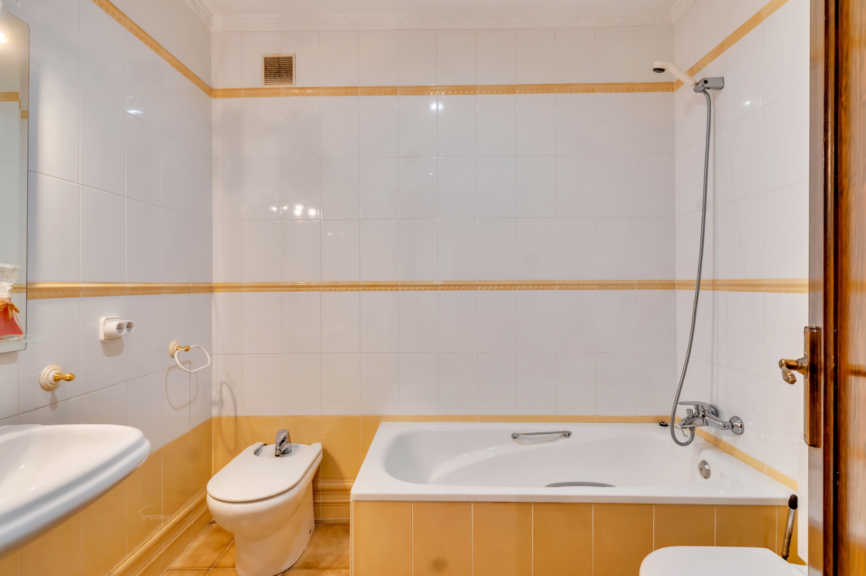 Cuarto de baño con azulejos blancos y detalles en ocre, equipado con bañera, bidet y grifería clásica. El diseño incluye molduras decorativas y accesorios en tonos dorados.