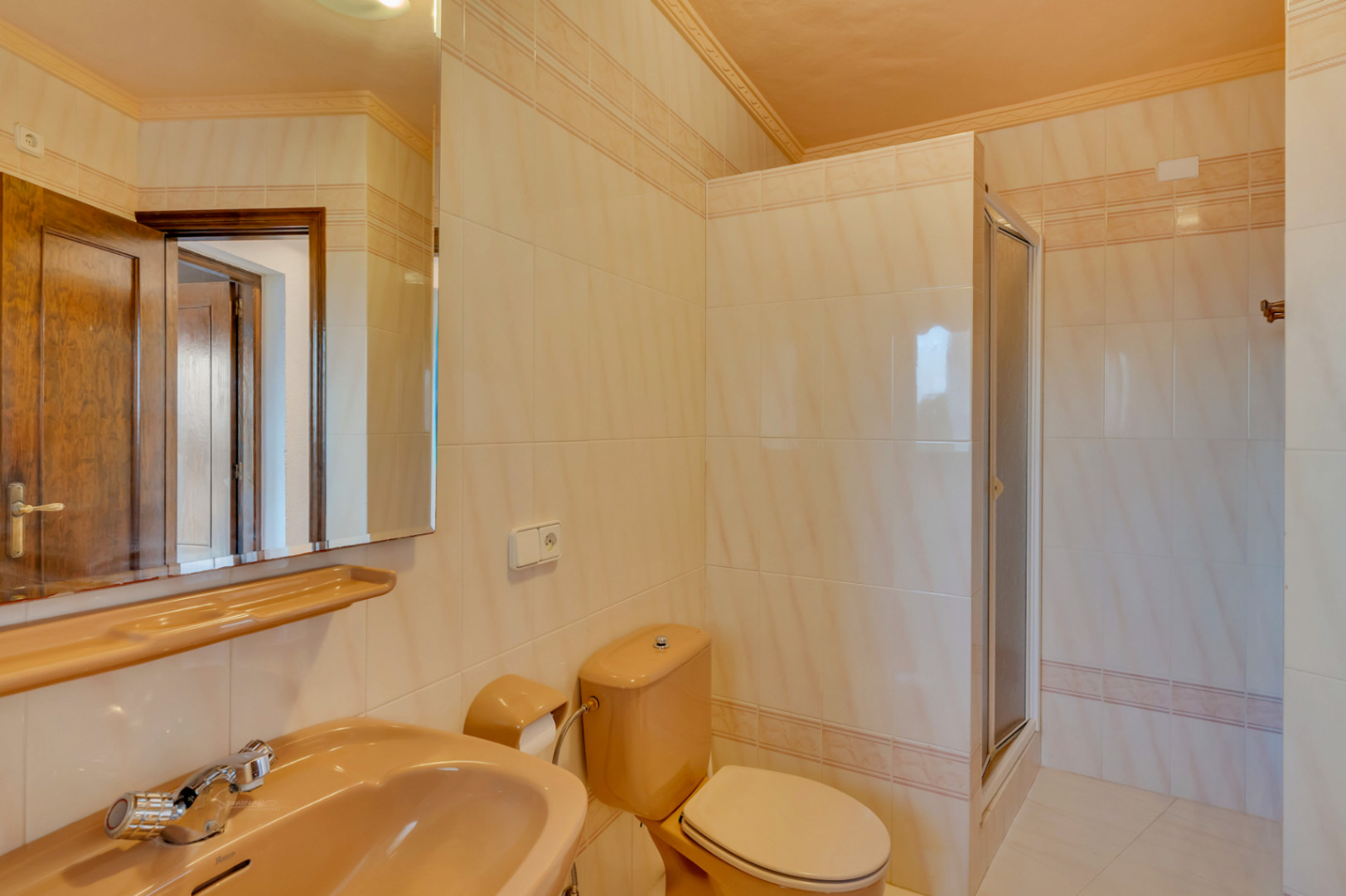 Cuarto de baño con azulejos en tonos crema, sanitarios de color melocotón y una ducha con mampara. Incluye un amplio espejo y acabados de estilo clásico.