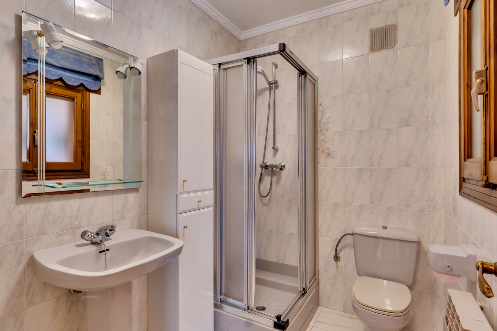 Cuarto de baño con lavabo de pedestal, espejo con iluminación integrada, armario blanco y cabina de ducha con mampara corredera.