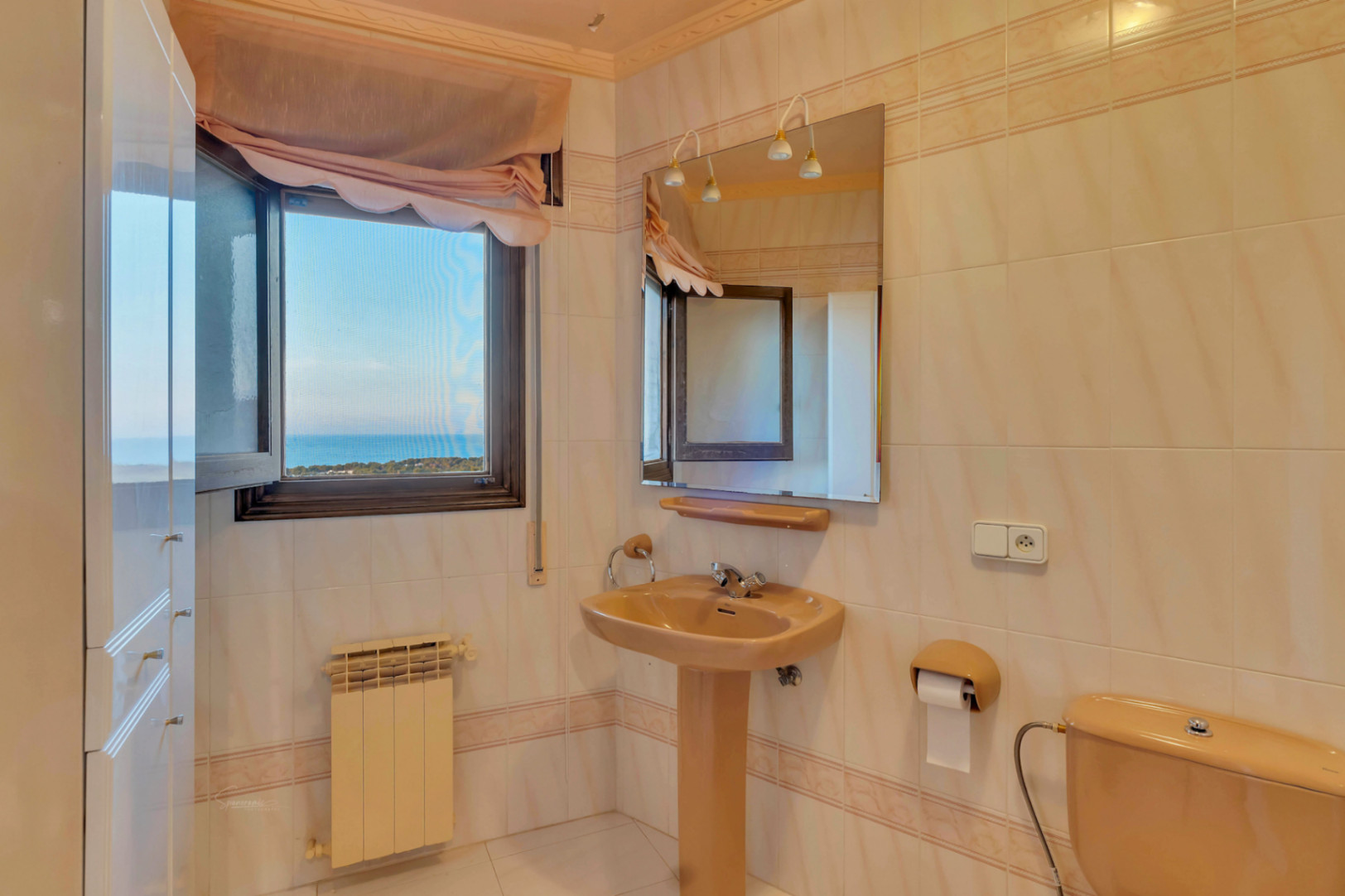 Cuarto de baño con sanitarios en color crema, lavabo de pedestal y ventana con vistas directas al mar Mediterráneo y la costa.