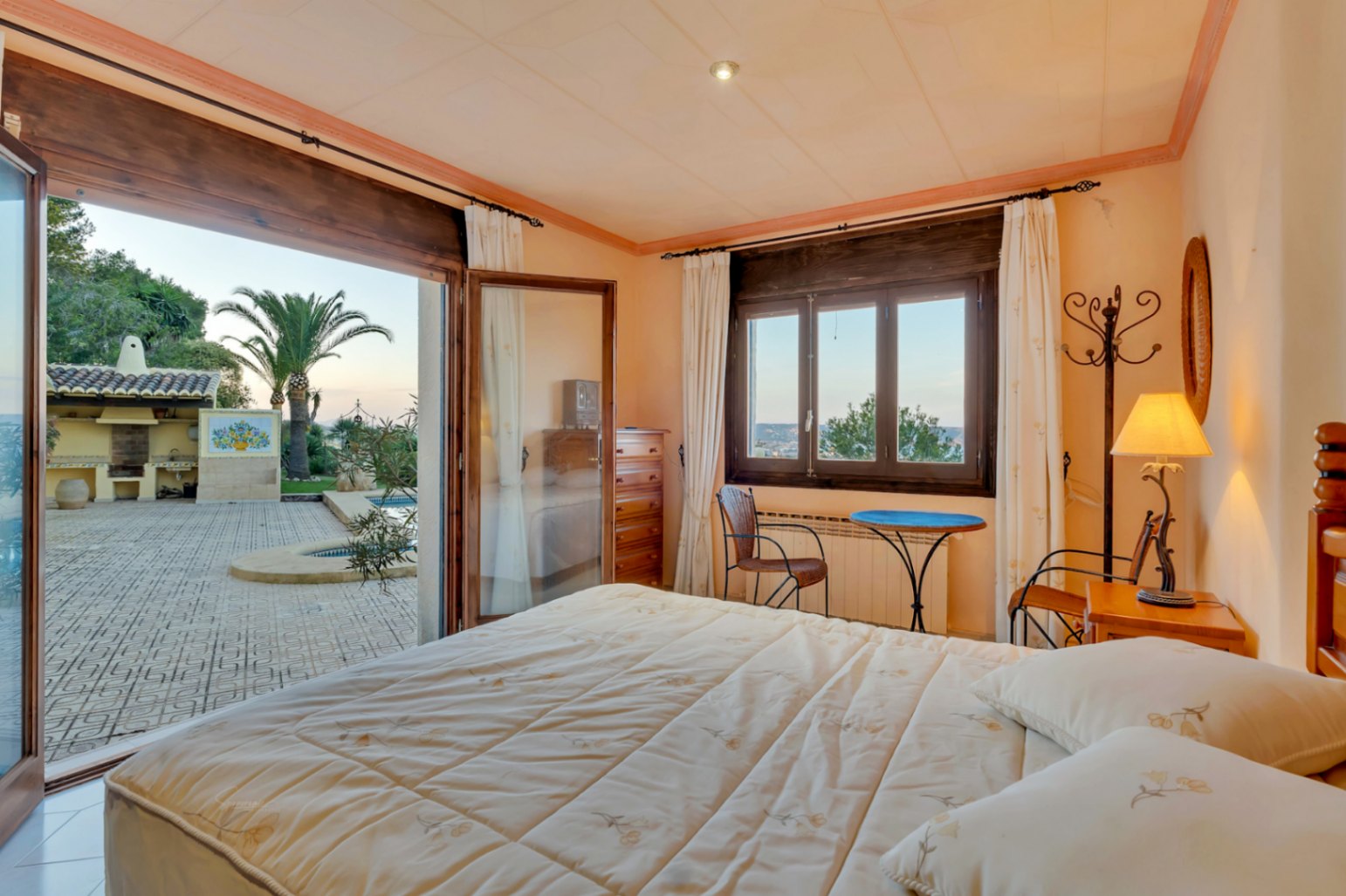 Dormitorio con acceso directo a la terraza y piscina, marcos de madera oscura, mobiliario clásico y vistas al jardín mediterráneo con zona de barbacoa.