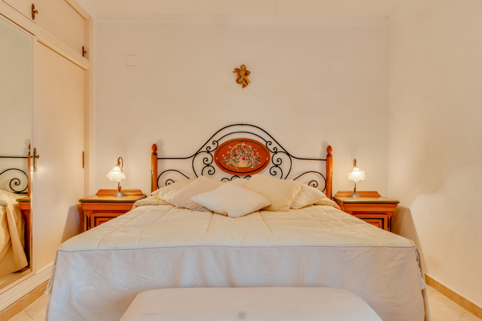Dormitorio con cama doble, cabecero de hierro forjado con medallón floral, mesitas de noche de madera y armario empotrado con espejo.