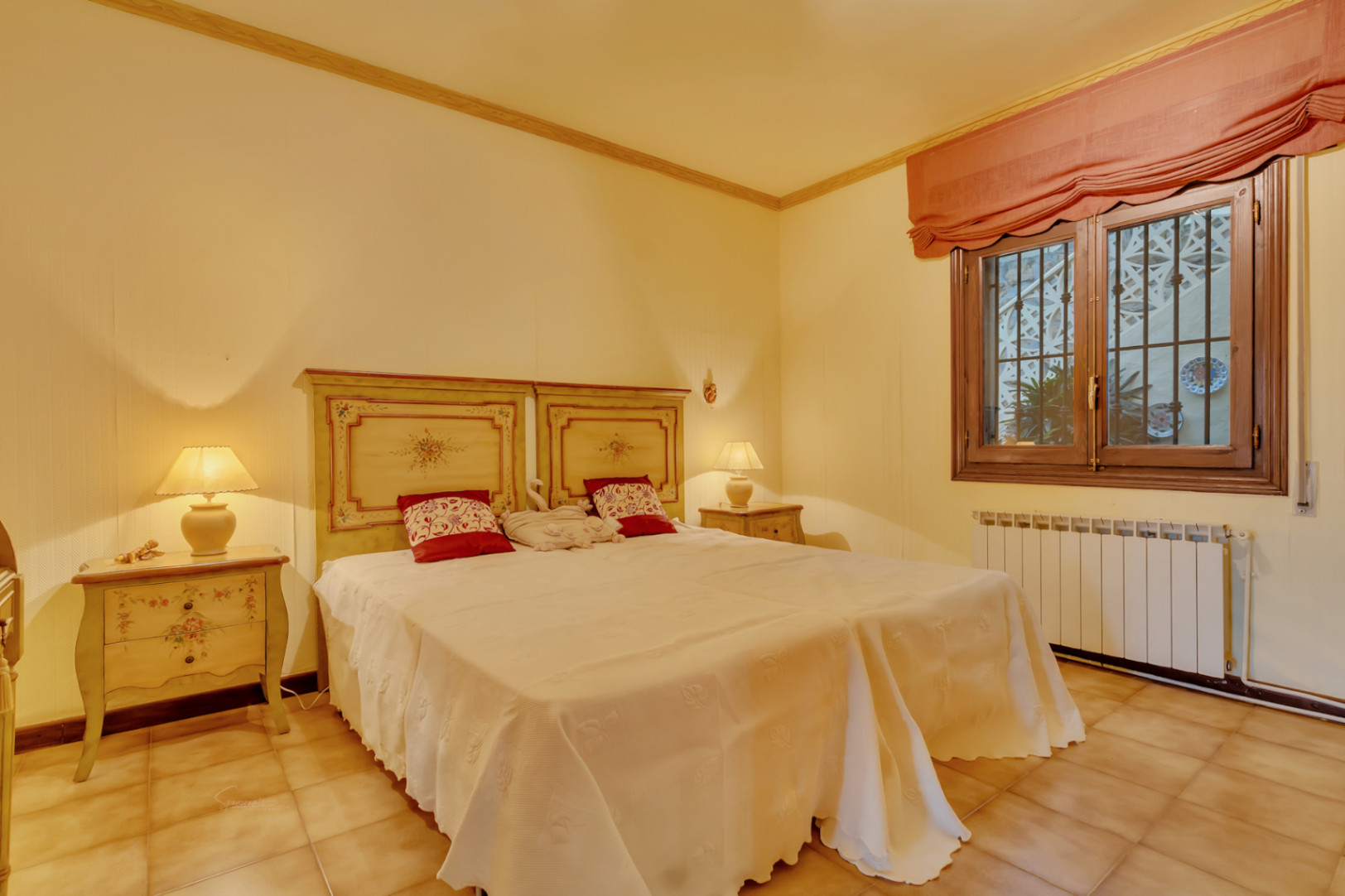 Dormitorio con camas gemelas, cabeceros de madera decorados, suelos de terracota, radiador de calefacción y ventana con marco de madera.