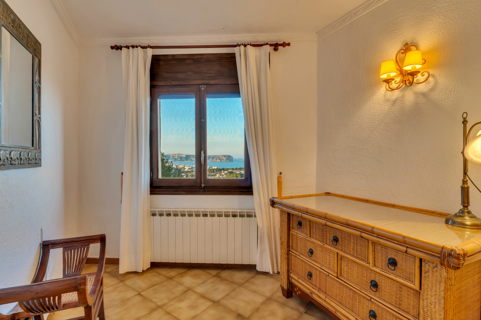 Dormitorio con cómoda de mimbre, suelos de gres y ventana de madera con vistas al Cabo San Antonio. Incluye calefacción por radiador y apliques de pared clásicos.