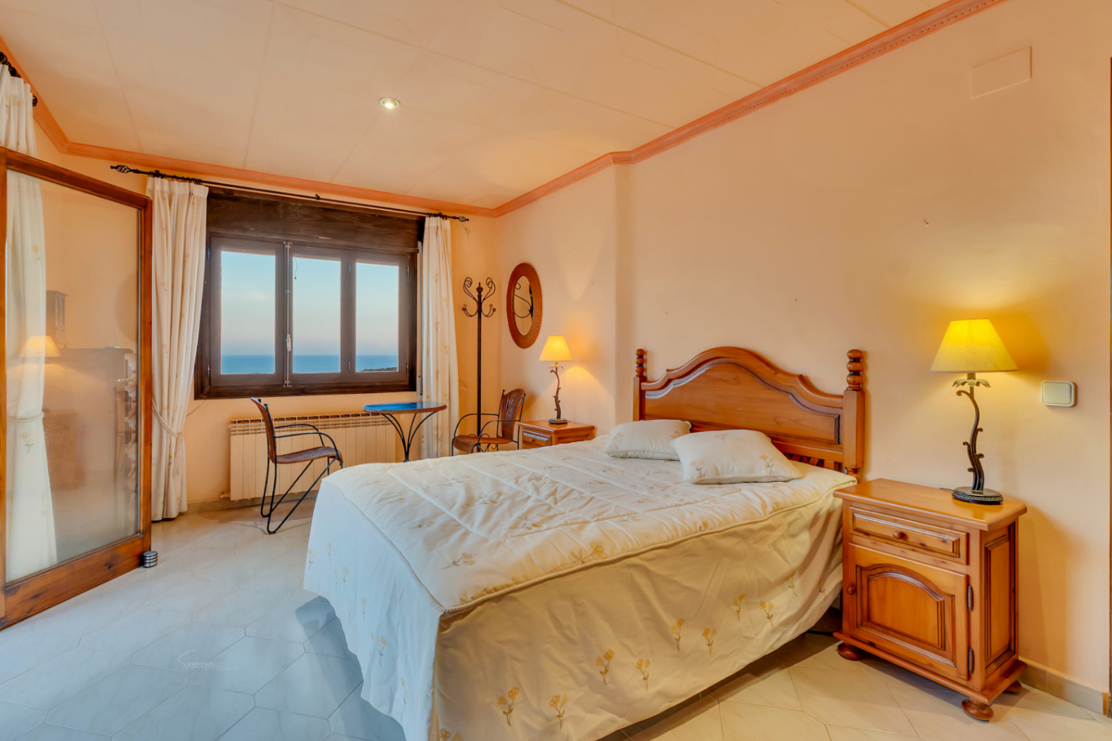 Dormitorio con mobiliario de madera clásica, suelos de baldosa hexagonal y ventana con vistas al mar. Incluye calefacción por radiador y una pequeña zona de estar.