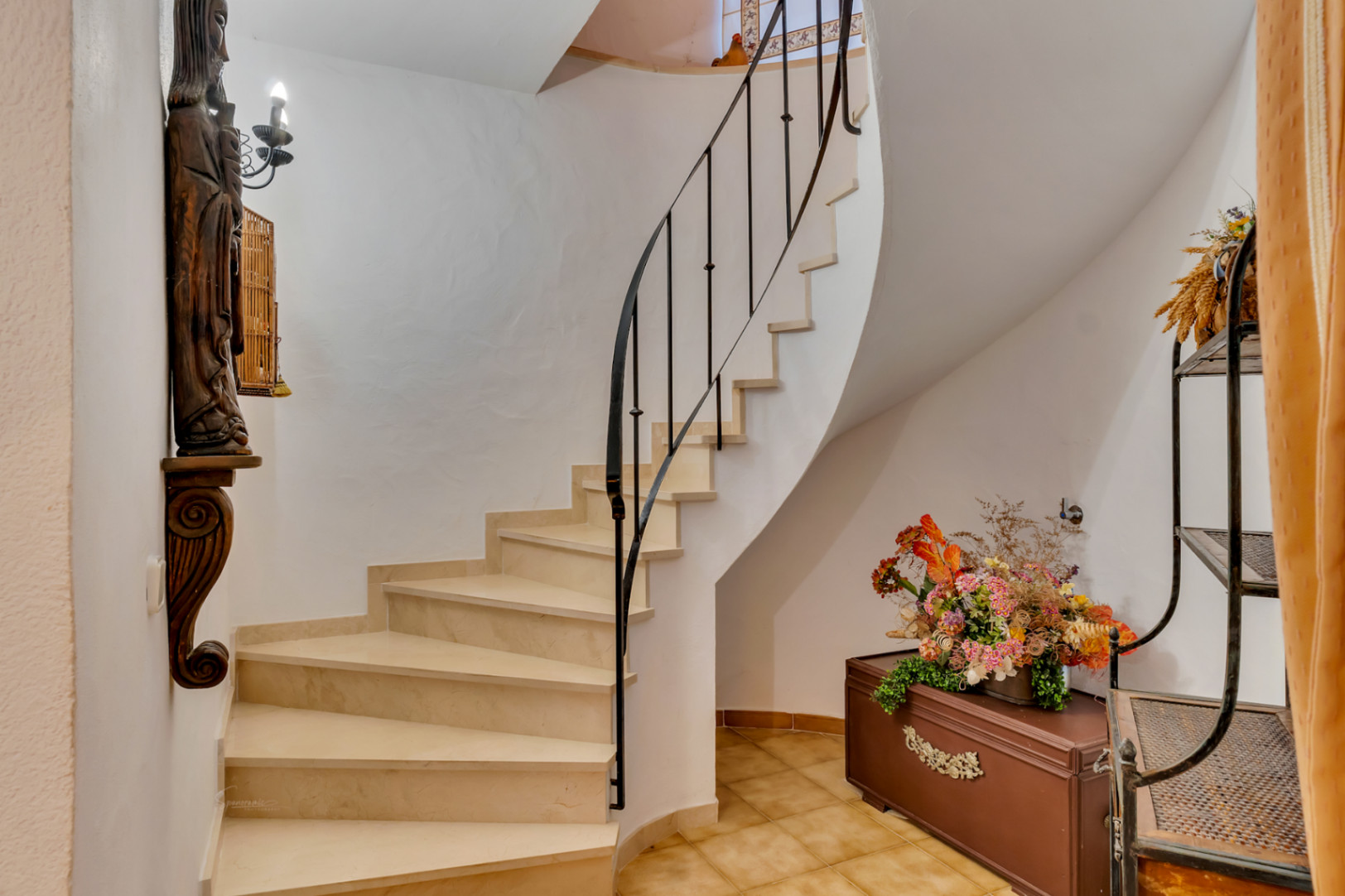 Escalera curva con peldaños de piedra clara y barandilla de hierro forjado negro. El espacio incluye paredes blancas y suelos de baldosa cerámica tradicional.