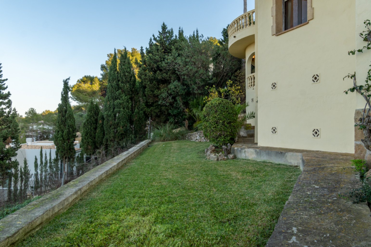Jardín mediterráneo con césped cuidado, rodeado de cipreses y vegetación autóctona. Se observa el lateral de la villa con balcón curvo y muros de piedra.